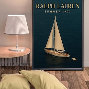 Op de afbeelding: Een ingelijste print met een zeilboot op een donkerblauwe zee. De tekst "RALPH LAUREN SUMMER 1997" staat in gouden letters. De zeilboot heeft witte zeilen en een houten romp. Een kleine boot is in de buurt zichtbaar.