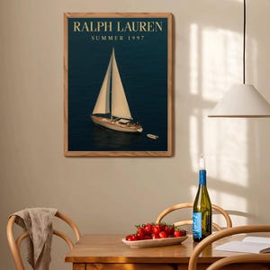 Op de afbeelding: Een ingelijste print met een zeilboot op donkerblauw water. De print heeft de tekst "RALPH LAUREN SUMMER 1997" in gouden letters. De lijst is gemaakt van lichtbruin hout. De afbeelding hangt aan een beige muur.