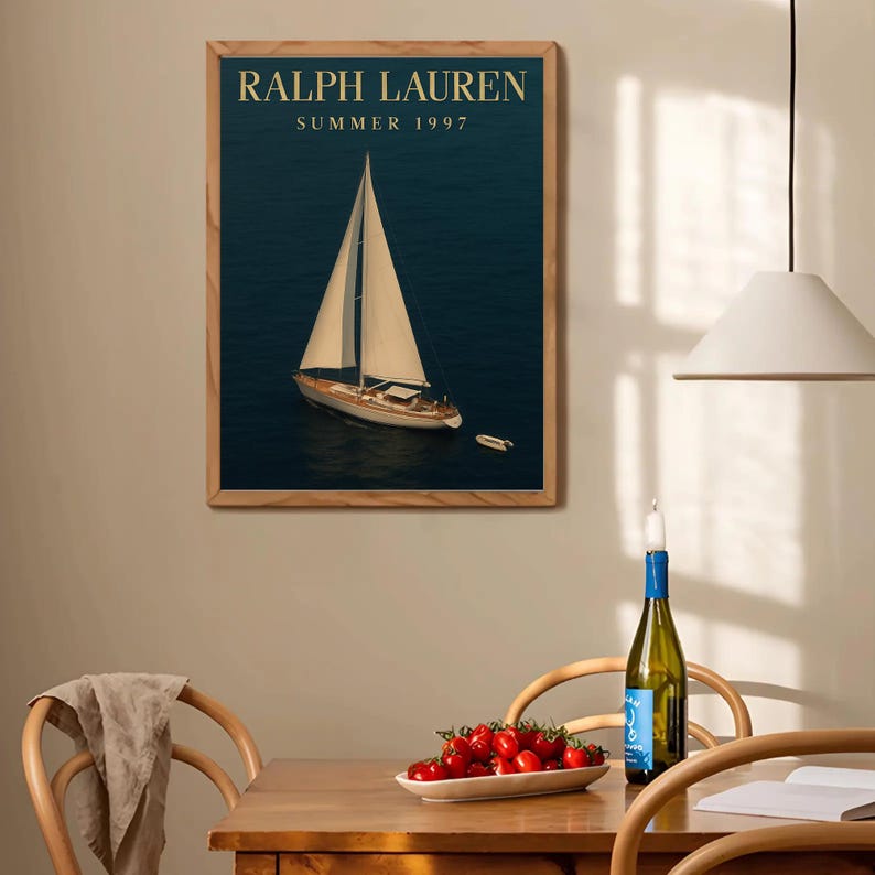 Op de afbeelding: Een ingelijste print met een zeilboot op donkerblauw water. De print heeft de tekst "RALPH LAUREN SUMMER 1997" in gouden letters. De lijst is gemaakt van lichtbruin hout. De afbeelding hangt aan een beige muur.