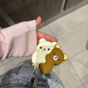 May include: A white and brown cartoon bear retractable badge holder. The badge holder features two bear figures hugging each other. The badge holder is attached to a silver metal clip.
