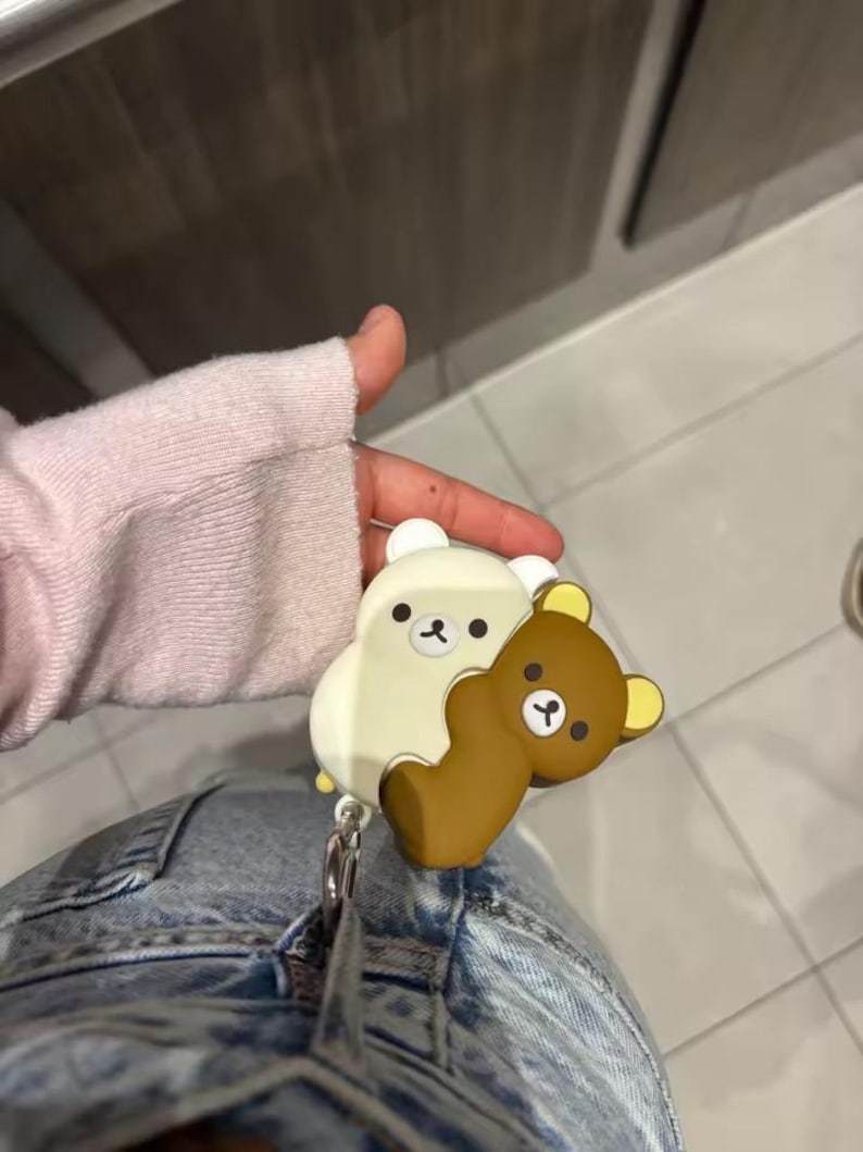 May include: A white and brown cartoon bear retractable badge holder. The badge holder features two bear figures hugging each other. The badge holder is attached to a silver metal clip.