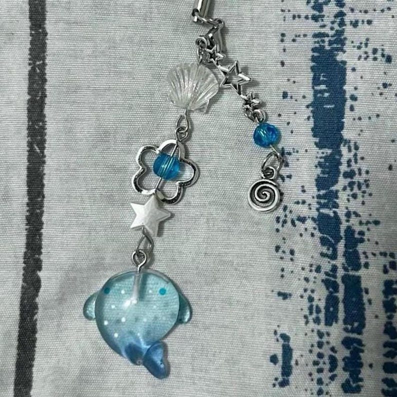 May include: A blue dolphin charm with silver star, flower, and seashell accents. The charm is attached to a silver chain with blue beads.