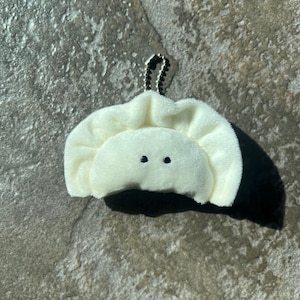 May include: A plush, off-white dumpling-shaped keychain with two small black eyes. The dumpling has a ruffled edge and a small silver chain for attachment. The background is a textured, gray stone surface.