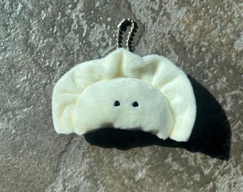 May include: A plush, off-white dumpling-shaped keychain with two small black eyes. The dumpling has a ruffled edge and a small silver chain for attachment. The background is a textured, gray stone surface.