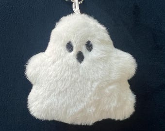 May include: A fluffy white ghost-shaped keychain with black eyes and mouth. The plush toy has a silver chain and clasp for easy attachment. The background is a dark blue.