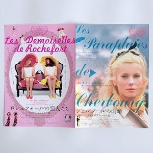 May include: A double-sided promotional print featuring movie posters for Les Demoiselles de Rochefort and Les Parapluies de Cherbourg. The left side is pink with two people on a sofa, while the right side features a portrait of a blonde person.