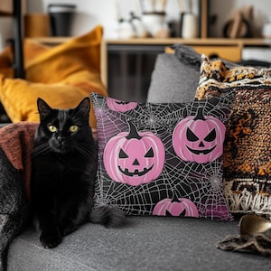 May include: A black cat sits on a gray couch with a pink and black throw pillow featuring a pattern of pink jack-o'-lantern faces and spiderwebs.