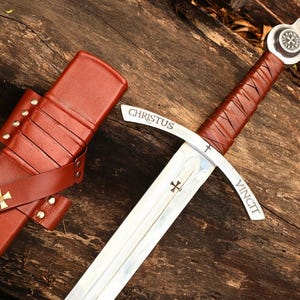 Pode incluir: Uma espada de estilo medieval com uma empunhadura envolta em couro castanho e uma lâmina prateada. A lâmina tem a inscrição "CHRISTUS VINCIT". A espada está ao lado de uma bainha de couro castanho com uma cruz dourada.