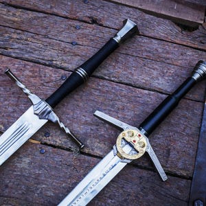 May include: Two medieval swords with silver blades and black handles are displayed on a weathered wooden surface. One sword has a decorative gold and red hilt. The swords have cross-shaped guards.