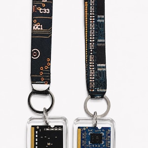 May include: Two keychains featuring fabric wrist straps with a black and copper circuit board pattern. Each strap is attached to a silver metal ring holding a square, clear acrylic pendant containing a small, rectangular computer memory module.