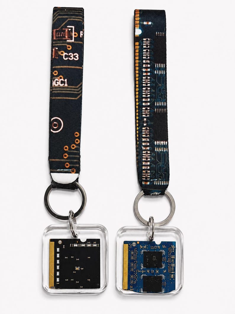 May include: Two keychains featuring fabric wrist straps with a black and copper circuit board pattern. Each strap is attached to a silver metal ring holding a square, clear acrylic pendant containing a small, rectangular computer memory module.