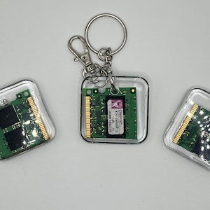 May include: Three clear square keychains with embedded green circuit boards. One keychain has a silver keyring and clasp. The circuit boards have gold and black components. The keychains are on a white surface.