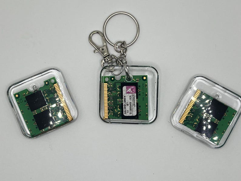 May include: Three square keychains featuring green computer RAM memory modules encased in clear acrylic. One keychain includes a silver metal split ring and lobster clasp. The central module displays a white label with the text Kingston, KTH-XW4300/1G, 1.8V, and ASME1840799.