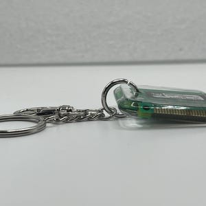 May include: A keychain featuring a rectangular piece of green computer circuit board encased in clear resin. The component is attached to a silver-toned metal split ring and a small chain link, showcasing the internal electronic pins and circuitry.