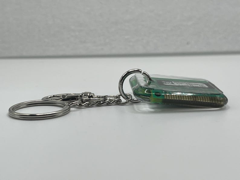 May include: A keychain featuring a rectangular piece of green computer circuit board encased in clear resin. The component is attached to a silver-toned metal split ring and a small chain link, showcasing the internal electronic pins and circuitry.