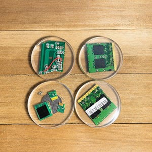 May include: Four circular clear coasters containing embedded green computer circuit board components. Each coaster features distinct electronic parts, including memory modules, small chips, and metallic connectors, all encased within a smooth, transparent resin material.