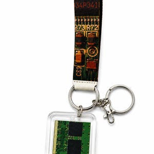May include: A keychain featuring a fabric strap printed with a green circuit board pattern and the text 9841 34P341 873 R72 C89. Attached to the strap is a silver metal ring and a clear square acrylic charm containing a green computer memory module.
