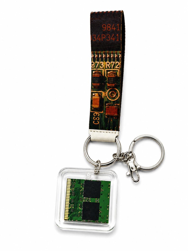 May include: A keychain featuring a fabric strap printed with a green circuit board pattern and the text 9841 34P341 873 R72 C89. Attached to the strap is a silver metal ring and a clear square acrylic charm containing a green computer memory module.