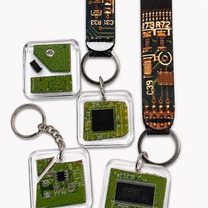 May include: A collection of four keychains featuring green circuit board fragments encased in clear square acrylic frames. Two keychains include long fabric straps printed with electronic circuit patterns, text, and codes like 984, 4P34, and C39.