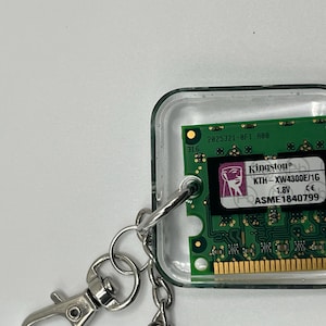 May include: A square keychain featuring a green computer RAM module encased in clear resin. The module displays the Kingston logo, model number KTH-XW4300E/1G, 1.8V, and serial number ASME1840799. A silver metal lobster clasp is attached to the side.