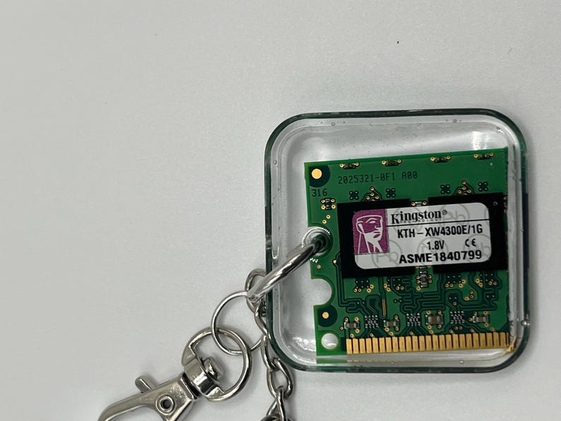 May include: A square keychain featuring a green computer RAM module encased in clear resin. The module displays the Kingston logo, model number KTH-XW4300E/1G, 1.8V, and serial number ASME1840799. A silver metal lobster clasp is attached to the side.