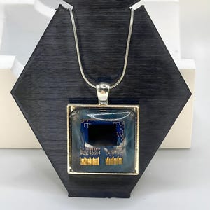 May include: A silver necklace with a square pendant featuring a circuit board design. The pendant has a gold-colored frame and a dark blue and black background. The necklace is displayed on a black stand.