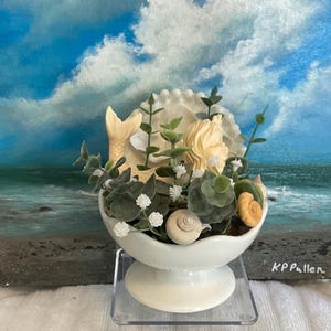 Puede incluir: Un cuenco de cerámica blanca con una figura de sirena y conchas marinas en su interior. El cuenco está sobre un soporte transparente y el fondo es una pintura de una escena de playa con cielo azul y nubes blancas.