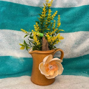 Pode incluir: Um pequeno jarro de cerâmica bege com uma alça, decorado com uma grande flor creme com um centro laranja. O jarro contém plantas artificiais amarelas e verdes e uma pequena estatueta de sapo verde.