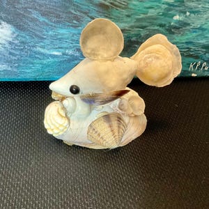 Puede incluir: Una escultura de ratón artesanal hecha con conchas marinas. El ratón está hecho de varias conchas en tonos blancos, crema y beige. Tiene un ojo de cuenta negro y una pequeña concha marrón para la nariz. Decoración costera única.