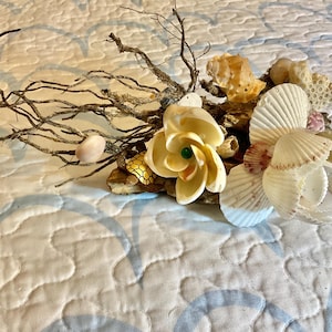 Pode incluir: Um arranjo floral de conchas com uma flor de concha branca e rosa, uma flor de concha branca com um centro verde e uma flor de concha branca com um centro rosa. O arranjo está sobre um pedaço de madeira à deriva com galhos e outras conchas.