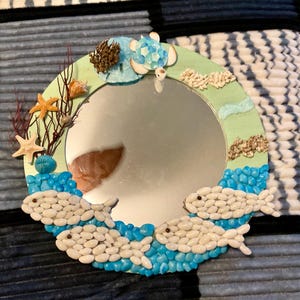 May include: Circular decorative mirror with a marine theme. The light green frame is decorated with seashells, starfish, and a turtle. Two fish made of white pebbles swim in a sea of blue pebbles, creating a coastal feel.