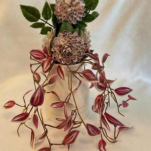 Może przedstawiać: Biały ceramiczny wazon z kompozycją kwiatową. Kompozycja zawiera duże, warstwowe, fioletowe kwiaty, zielone liście i pnącza z bordowymi i białymi liśćmi. Wazon stoi na białej powierzchni.