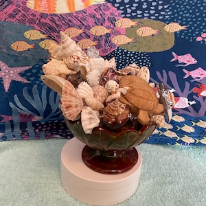 Pode incluir: Uma tigela de cerâmica com um esmalte verde e marrom, cheia de uma variedade de conchas e uma pequena tartaruga de madeira. A tigela está sentada em um pedestal branco.