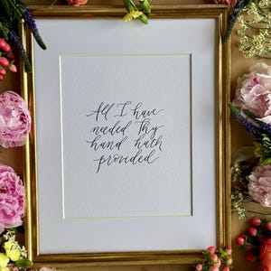May include: A framed print with the handwritten quote, "All I have needed Thy hand hath provided." The gold frame surrounds a white mat and the print. The frame is surrounded by colorful flowers, including pink and orange peonies.