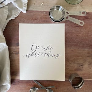 May include: A still-life image featuring a wooden surface with a white card inscribed with "Do the next thing" in elegant calligraphy. Measuring cups, spoons, a towel, and a glass vase are arranged around the card, suggesting a kitchen or craft theme.