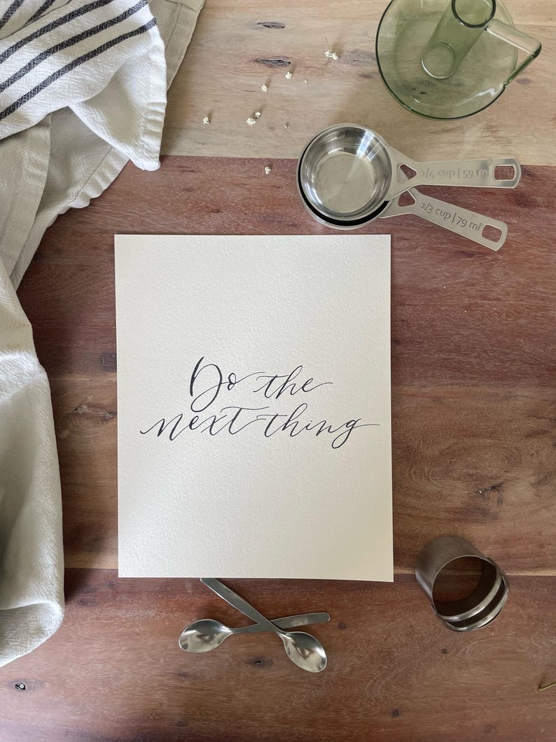 May include: A still-life image featuring a wooden surface with a white card inscribed with "Do the next thing" in elegant calligraphy. Measuring cups, spoons, a towel, and a glass vase are arranged around the card, suggesting a kitchen or craft theme.