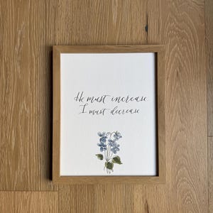 May include: A rectangular light wood frame containing a white print with the black handwritten text He must increase. I must decrease. Below the text is a small watercolor illustration of blue violet flowers with green leaves on a white background.