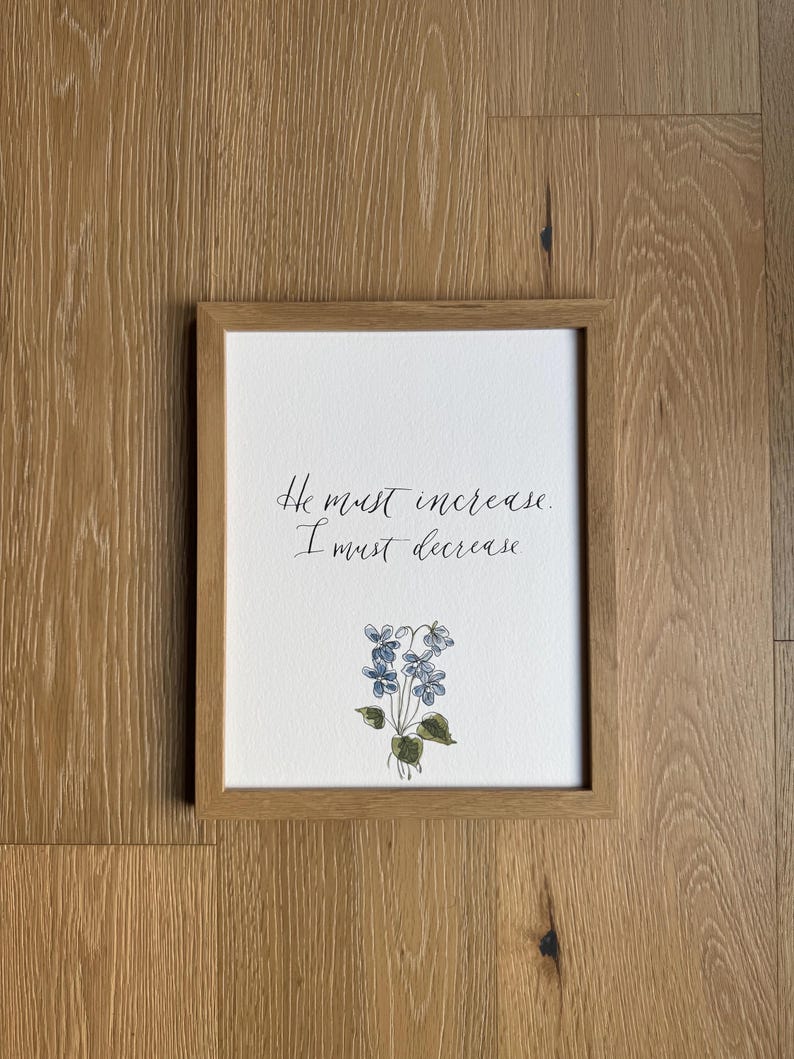 May include: A rectangular light wood frame containing a white print with the black handwritten text He must increase. I must decrease. Below the text is a small watercolor illustration of blue violet flowers with green leaves on a white background.
