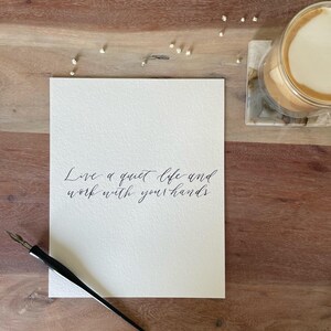 May include: A white card with the handwritten quote, "Live a quiet life and work with your hands." A black calligraphy pen rests on the card. A glass of coffee with foam sits on a coaster, and small white flowers are scattered on a wooden surface.