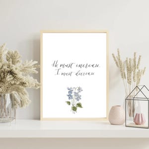 May include: A framed wall art print featuring the black cursive text He must increase. I must decrease. centered above a delicate watercolor illustration of a small cluster of blue violet flowers with green leaves on a white background.