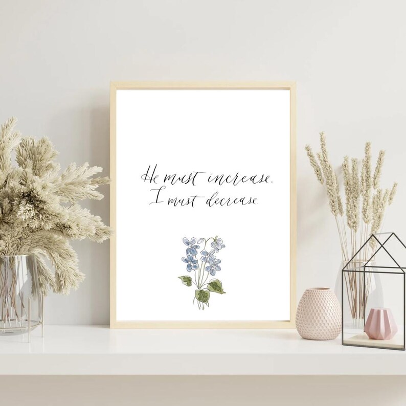 May include: A framed wall art print featuring the black cursive text He must increase. I must decrease. centered above a delicate watercolor illustration of a small cluster of blue violet flowers with green leaves on a white background.
