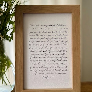 May include: A framed print with handwritten calligraphy in black ink. The wooden frame surrounds a white paper with text from Psalm 23. The print is displayed on a wooden surface, with flowers in a vase in the background.