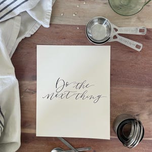 May include: A still-life image featuring a wooden surface with various kitchen tools and a card. The card has the phrase "Do the next thing" in elegant script. Measuring cups, spoons, and a pastry cutter are also visible.