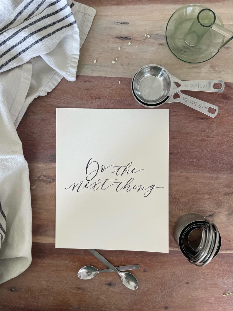 May include: A still-life image featuring a wooden surface with various kitchen tools and a card. The card has the phrase "Do the next thing" in elegant script. Measuring cups, spoons, and a pastry cutter are also visible.