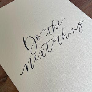 May include: A cream-colored paper with the handwritten phrase "Do the next thing" in black calligraphy. The paper has a textured surface, and the text is elegantly scripted.