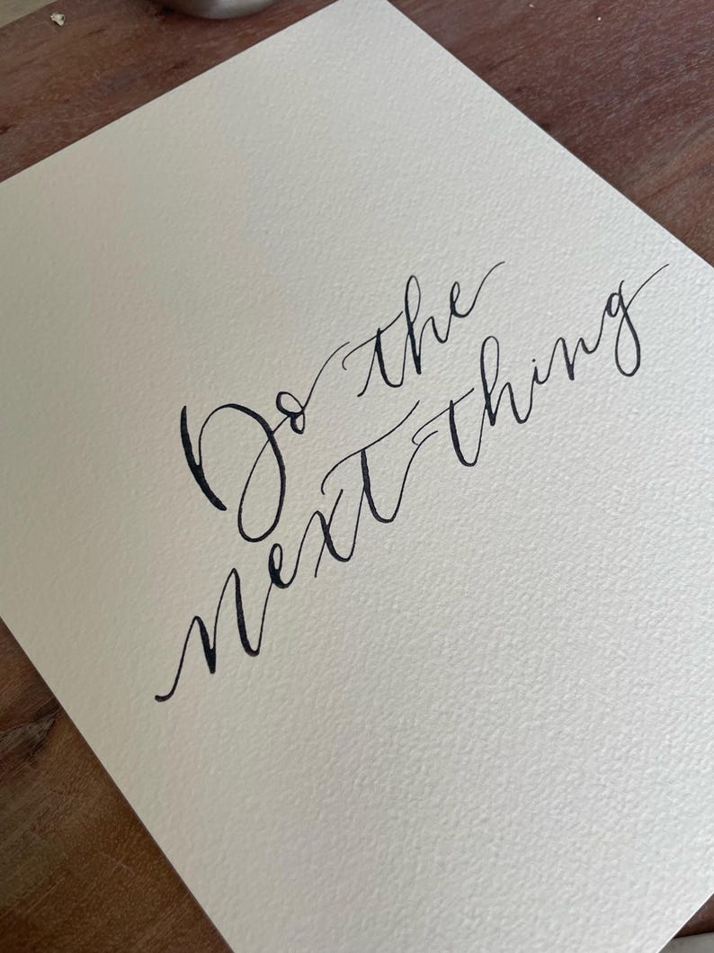May include: A cream-colored paper with the handwritten phrase "Do the next thing" in black calligraphy. The paper has a textured surface, and the text is elegantly scripted.
