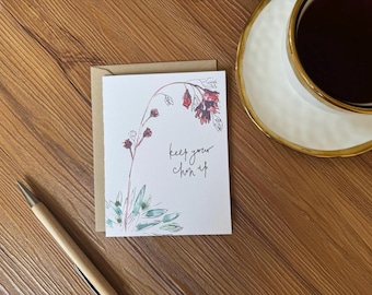 May include: A greeting card with a floral illustration and the handwritten phrase "keep your chin up." The card is on a wooden surface, next to a pen and a cup of coffee.