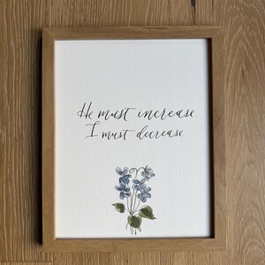 May include: A framed print featuring the black handwritten text "He must increase. I must decrease." centered above a delicate watercolor illustration of a small cluster of blue violet flowers with green leaves, enclosed in a light brown wooden frame.