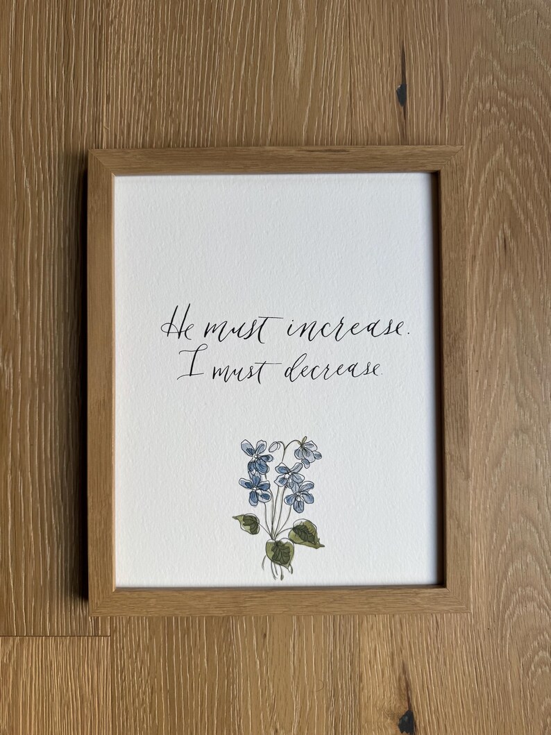 May include: A framed print featuring the black handwritten text "He must increase. I must decrease." centered above a delicate watercolor illustration of a small cluster of blue violet flowers with green leaves, enclosed in a light brown wooden frame.