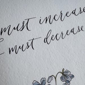 May include: A white textured paper featuring black calligraphy text that reads "must increase" and "I must decrease" positioned above a hand-drawn illustration of small blue flowers with green stems and leaves rendered in a watercolor style.
